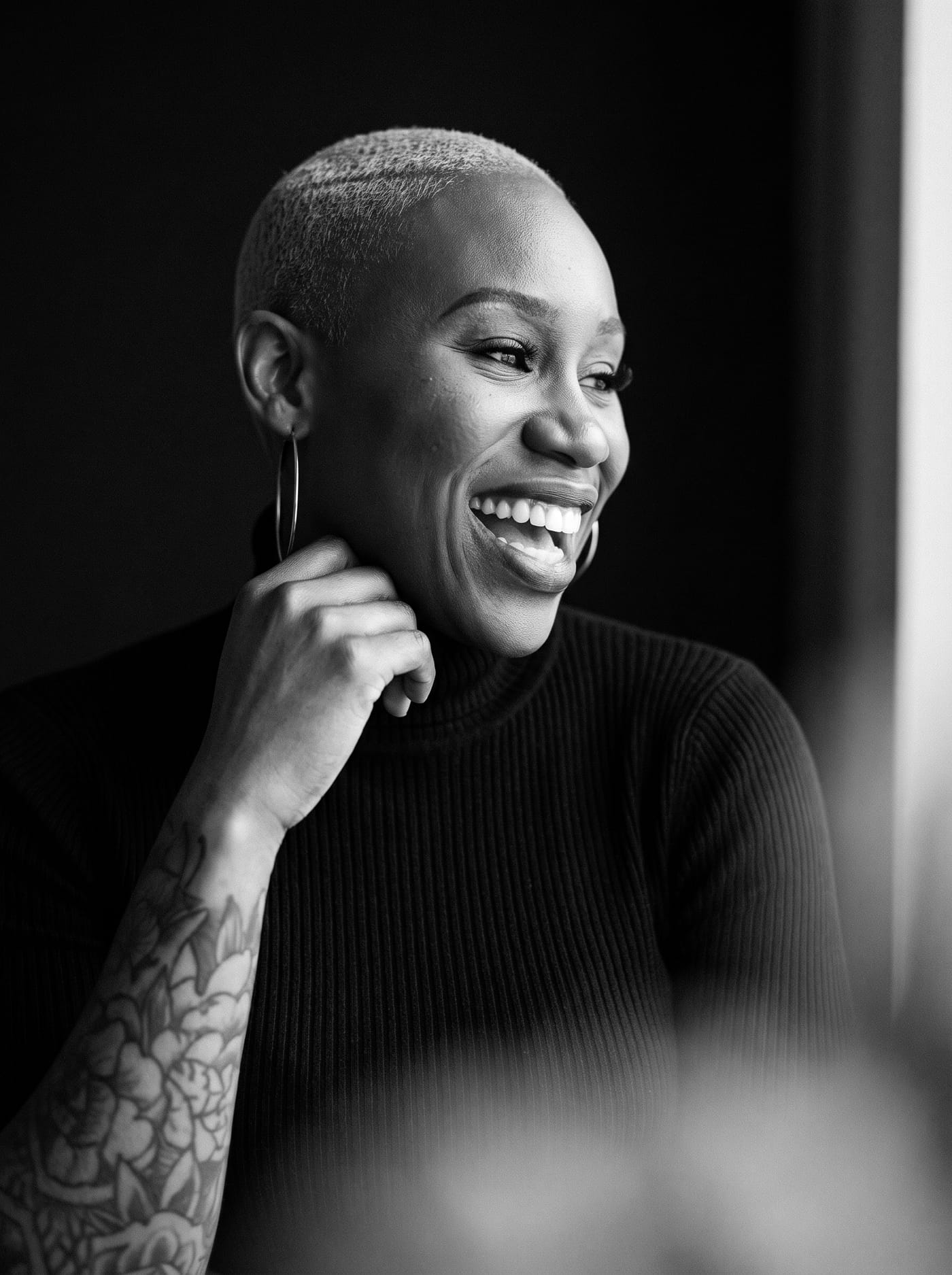Black and white candid portrait of Nikki Rumph laughing in a black turtleneck with her tattooed arm raised