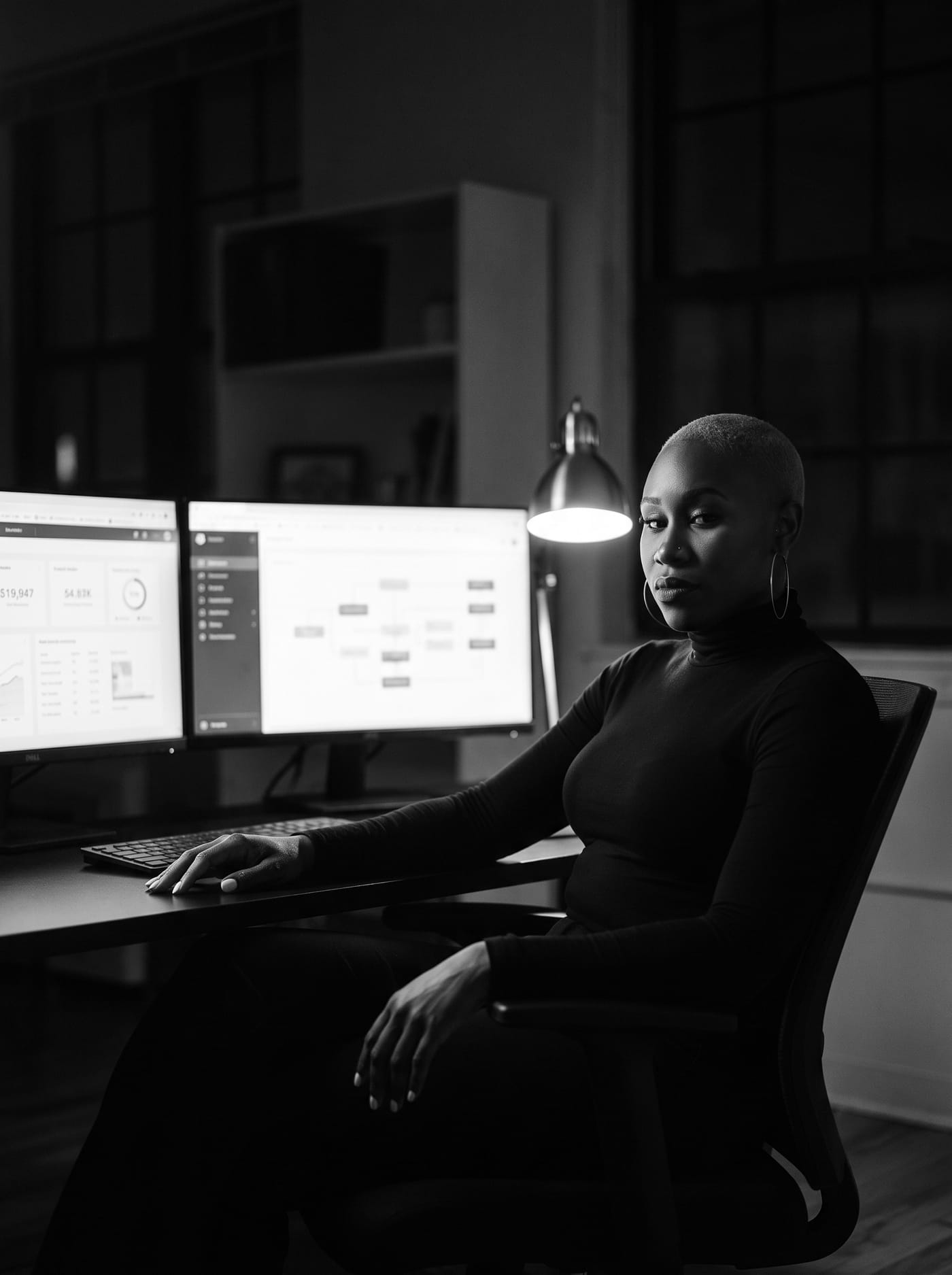 Black and white portrait of Nikki Rumph seated at a desk with two monitors showing dashboards under a desk lamp