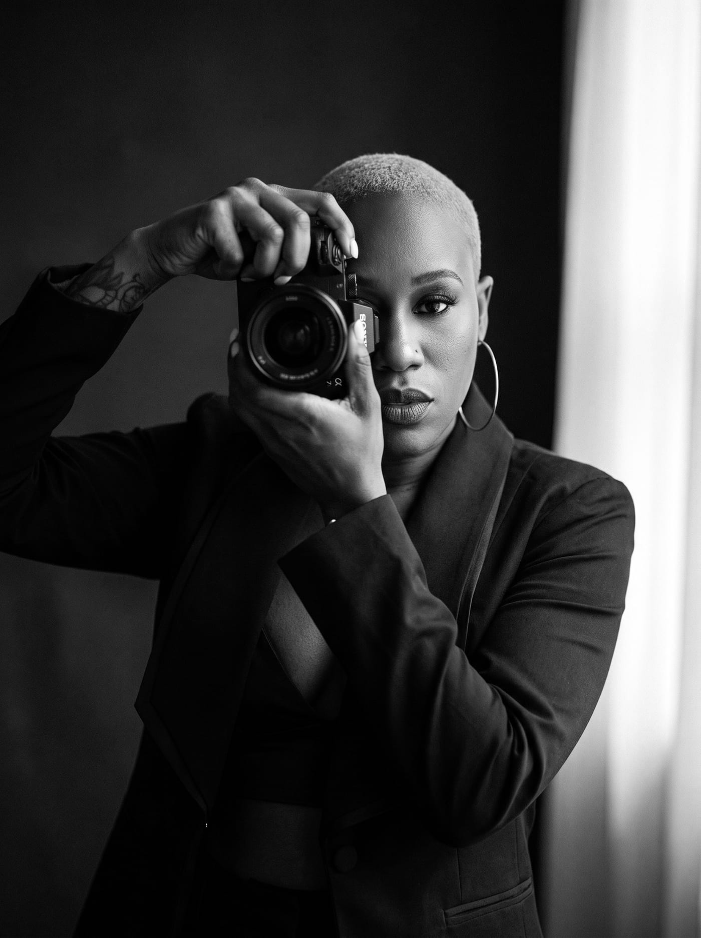 Black and white portrait of Nikki Rumph raising a camera to her eye beside a bright window