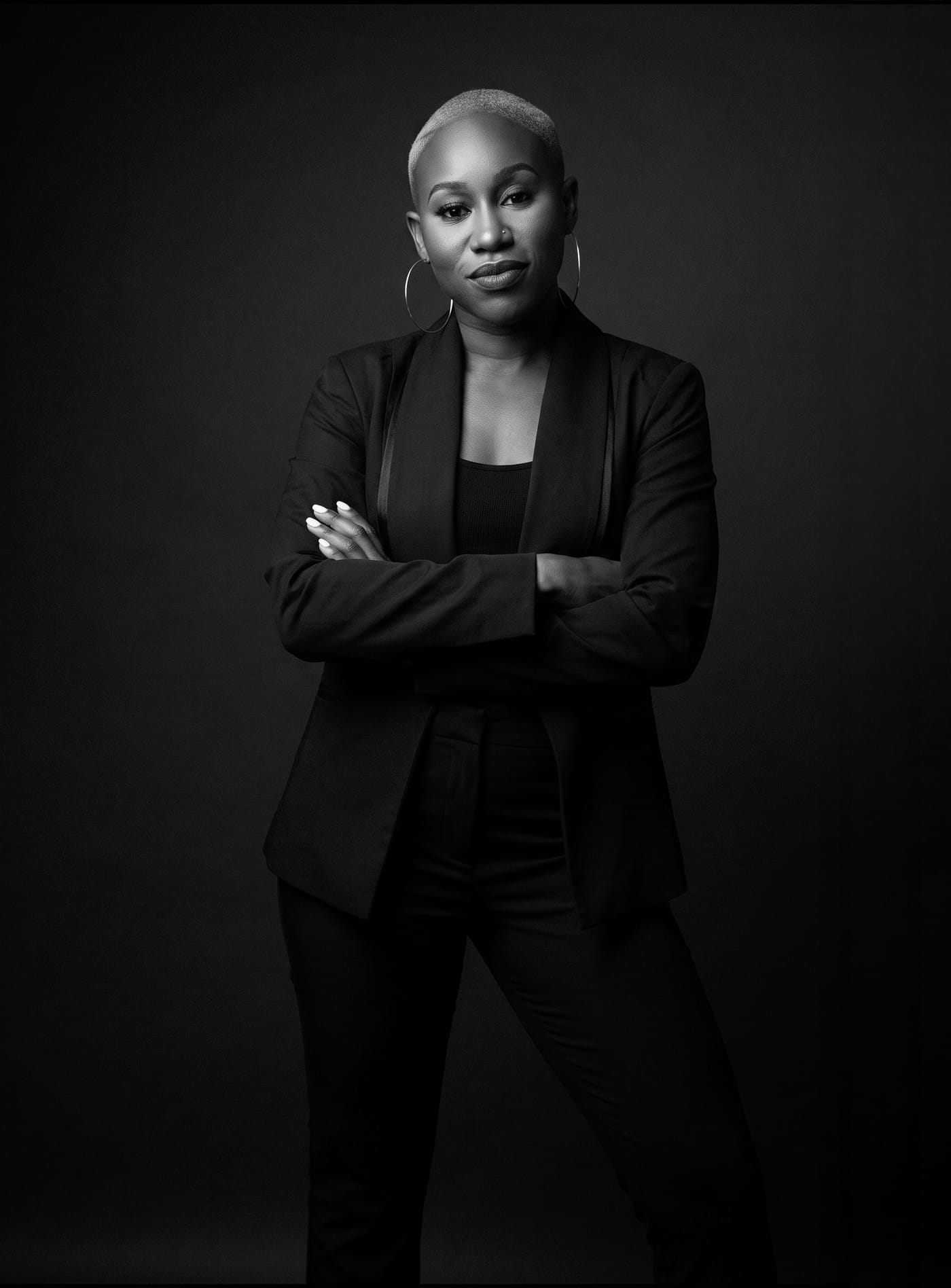 Black and white portrait of Nikki Rumph standing with her arms crossed in an all-black blazer against a dark studio backdrop